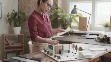 Modern tavan arası ofisindeki çalışma masasında duran ve 3D modeline ve planlarına bakarak ev tasarlayan kadın mimar yavaşlıyor.