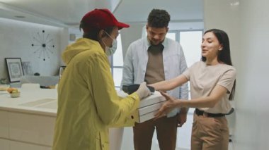 Sarı iş kıyafetleri ve yüz maskesi takmış orta boy erkek kuryenin parlak, modern bir dairede yaşayan genç Latin çifte üç büyük pizza kutusu getirmesi. Hesabı kredi kartıyla ödeyen adam.