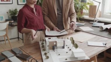 Yeni ev tasarımı taslaklarını inceleyen profesyonel mimarlardan oluşan eğik-büğrü bir ortam, çalışma masasındaki 3D düzeniyle karşılaştırıyor.