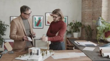 Modern ev için inşaat malzemelerini tartışan profesyonel erkek mimarların yavaş çekimleri modern çatı katındaki çalışma masasının 3D düzenine bakıyor.
