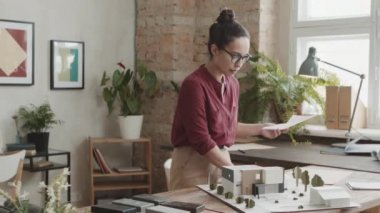 Profesyonel kadın mimarın orta PAN yavaşlığı modern ev tasarımı yaratıyor. Çalışma masasının üzerindeki taslaklar ve 3D düzen parlak çatı katı tarzı ofis tarzında.
