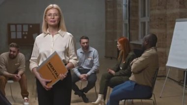 Kendine güvenen kadın psikoloğun orta PAN portresi zihinsel destek grubu başlığı ile klasörü tutarken grup toplantısına katılanlar arka planda konuşurken kamera için poz veriyor