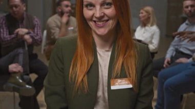 Ofis kıyafetleri içinde gülümseyen kızıl saçlı kadının yakın plan portresi ve diğer üyeler arka planda sohbet ederken grup terapisinde kameraya gülümseyen isim etiketi.