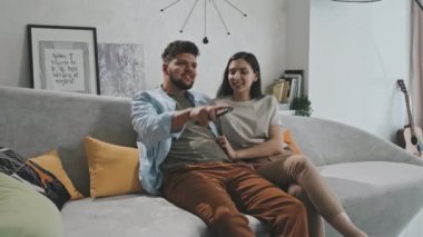 Modern oturma odasındaki kanepede oturan ve TV 'de komedi izlerken birlikte film izledikleri mutlu, genç bir Latin çiftin orta boy PAN çekimi.