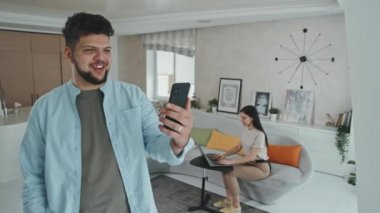 Genç Latin bir adamın akıllı telefondan arkadaşlarıyla ya da akrabalarıyla yaptığı konuşmanın orta boy görüntüsü. Karısı ise rahat, modern oturma odasındaki kanepede otururken.