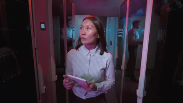 データセンターをタブレットで歩くアジアの女性技術者の撮影を追跡し 青いケーブルでスイッチやその他のコンピューティング機器をチェックする — ストック動画
