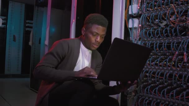 非洲裔美国男性网络工程师蹲伏在装有蓝色电缆的交换机前并在笔记本电脑上打字的中景照片 — 图库视频影像