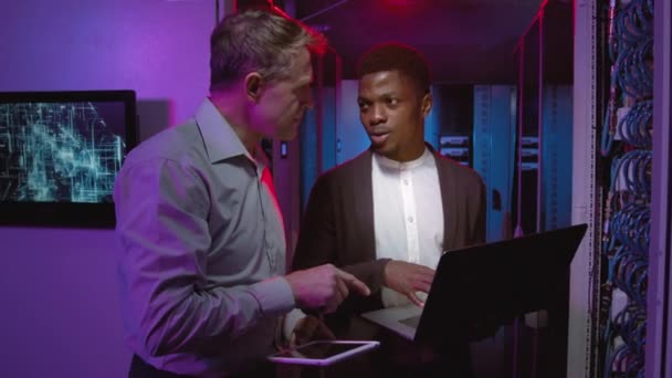 中年男性の追跡ショット白人ネットワークエンジニアと彼の男性アフリカ系アメリカ人の同僚ノートパソコンやタブレットを保持し データセンターでサーバーやスイッチをチェックしながら話す — ストック動画