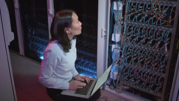 データセンター内のスイッチや他のコンピューティング機器の前にうずくまって ラップトップ上で入力する女性アジアのネットワークエンジニアの追跡ショット — ストック動画