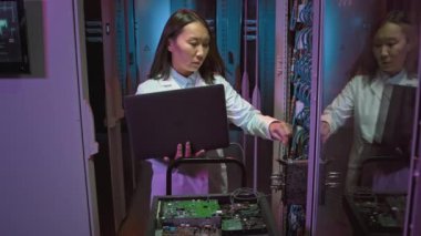 Dişi Asyalı bilişim uzmanının laptopunu veri merkezindeki bilgisayar ekipmanına bağlayan ve bir şeyi kontrol eden laboratuvar önlüğü üzerindeki yavaşlığını izliyorum.