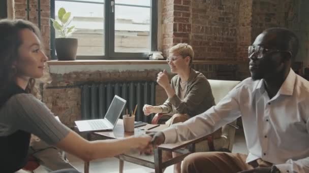 アフリカ系アメリカ人のビジネスパートナーに笑顔を見せる若い魅力的なビジネス女性たちが握手を交わしながら 同僚たちが背景で会話をしながら — ストック動画