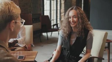 Orta yaşlı, çekici kıvırcık saçlı, uzun kıvırcık saçlı bir kadın, bayan meslektaşıyla konuşurken gülümsüyor ve modern çatı katında birbirlerine beşlik çakıyor.