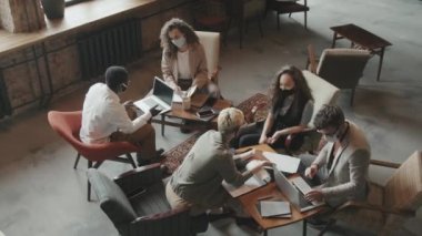 Üst görüşlü çok ırklı genç iş ortaklarının yüz maskeleri altında tavan arası iş yerinde beyin fırtınası yaparken yavaşlaması ve proje başarısı sonrasında birbirlerine beşlik çakmaları