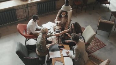 Çağdaş çatı katında çalışma maskesinde oturan ve sonra da takım çalışması ve ortaklık kavramının ortasında el ele tutuşan koruyucu maskeler takan genç iş arkadaşlarından oluşan üst düzey yavaşlama.