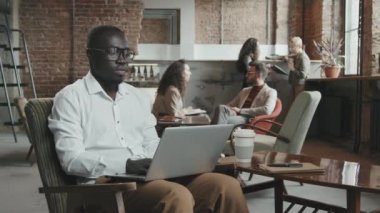 İş arkadaşları arka planda sohbet ederken şık beyaz tişörtlü ve gözlüklü, rahat tavan arası çalışma alanında dizüstü bilgisayarla çalışan, konsantre, genç, Afro-Amerikalı bir adam.