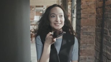 Modern çatı katının koridorunda kameraya doğru yürüyen ve akıllı telefondan sesli mesaj kaydeden neşeli, genç ve neşeli yarış işkadını.