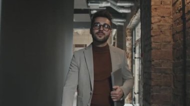Gözlüklü genç ve yakışıklı işadamının dizüstü bilgisayarla kameraya doğru yürüdüğü orta boy bir yavaşlama. Çağdaş çatı katı ofis koridorunda.