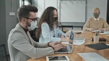 Yüzü maskeli ciddi genç işadamlarının iş yerinde kadın meslektaşlarıyla birlikte kameraya bakması ve dizüstü bilgisayarla çalışması.