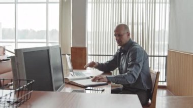 Orta yaşlı bir erkek profesörün modern parlak sınıftaki masasında oturup dizüstü bilgisayarla çevrimiçi ders almasının yan görüntüsü.