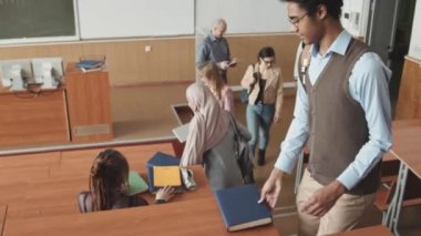 Yüksek açılı, çok ırklı bir grup genç öğrencinin ders başlamadan önce birbirlerini selamladıkları konferans salonundaki uzun tahta masalarda yerlerini almalarının yavaş çekimi.
