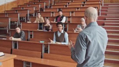 Büyük konferans salonu veya konferans salonunda ayakta duran çok ırklı küçük bir grup öğrenciye yeni materyalleri açıklayan kendinden emin erkek üniversite profesörünün orta derece yavaşlaması