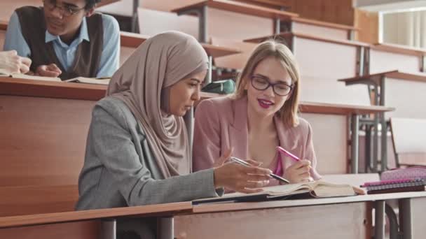 两个年轻的多民族女生坐在大学礼堂的课桌边 一边看书 一边说着话 — 图库视频影像