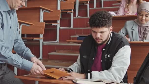 中等速度慢的身穿运动轰炸机夹克的年轻拉丁大胡子学生看着中年教授在考试或考试中帮助他的抄本中的笔记 — 图库视频影像