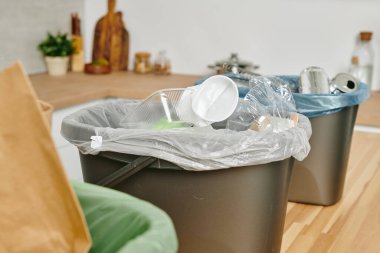 Kullanılmış tek kullanımlık mutfak gereçleri ve ahşap masadaki çöp kutusunda diğer atıklarla dolu plastik bir torba.