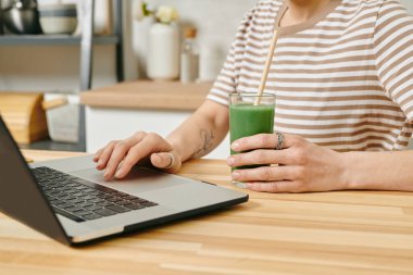 Genç bir bayan içki tutuyor ve masa başı dizüstü bilgisayar kullanıyor.