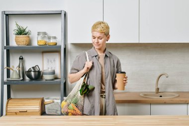 Elinde bir torba taze sebze ve kağıt bardak kahve tutan genç bir kadın.