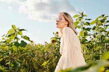 Beyaz elbiseli güzel sarışın kadın elinde bir şişe ayçiçeği yağı tutarken kameranın önünde büyük çiçekler arasında dikiliyordu.