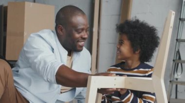 Mutlu Afro-Amerikan baba ve oğlunun yavaş yavaş yaklaşması ve yeni dairede tahta sandalye monte ederken yerde oturup sohbet etmeleri.