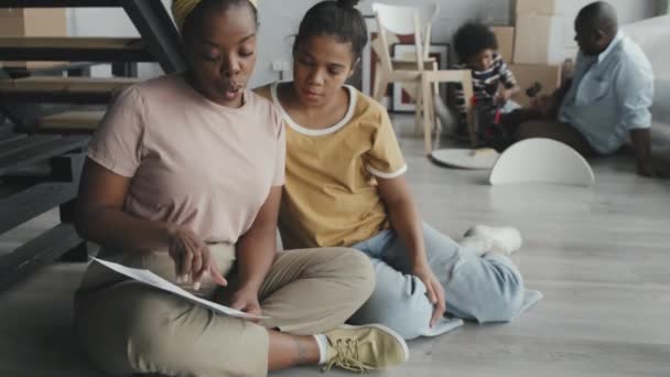 非洲裔美国妇女和女孩一起坐在地板上 看着组装说明 而父亲和孩子则在后面聊天和使用工具 — 图库视频影像