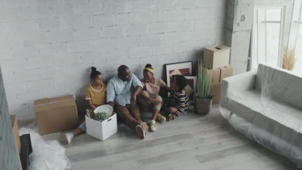 アフリカ系アメリカ人の家族の高角度のショット4つの床に新しいアパートの壁に座って移動した後 チャット彼らの持ち物は段ボール箱に入っています — ストック動画