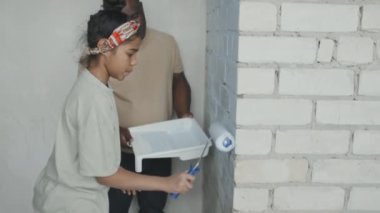 Afrika kökenli Amerikalı bir babanın elinde tepsiyle kızının tuğla duvarı boyadığını yavaş yavaş gösteren bir kare.