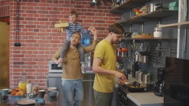 手持中镜头的快乐男人背着可爱的儿子走到他的男伴身边 在厨房做早餐 — 图库视频影像