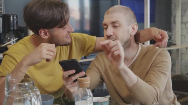 手握慢镜头的快乐同性恋夫妇坐在厨房桌子边一边聊天一边看手机 — 图库视频影像