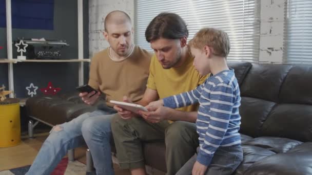 一对同性恋夫妇和他们的小儿子坐在沙发上 用平板电脑和手机拍了照 — 图库视频影像