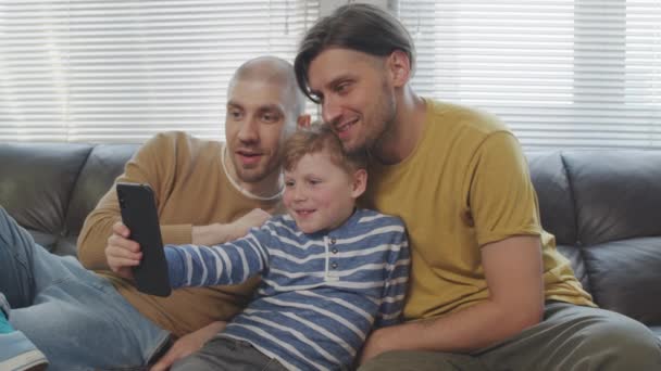 幸せなゲイの父親とかわいい息子の遅いショット一緒にソファに座って携帯電話で写真を撮る — ストック動画