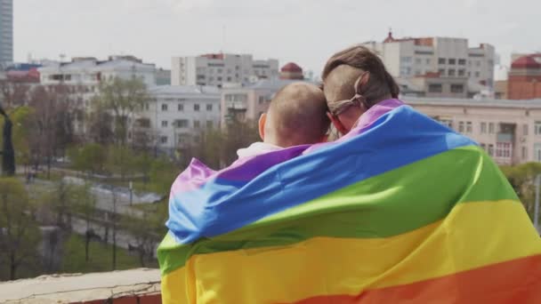 晴朗的夏日 一对手持式后视镜的同性恋夫妇抱着彩虹旗拥抱着屋顶 看城市风景 — 图库视频影像