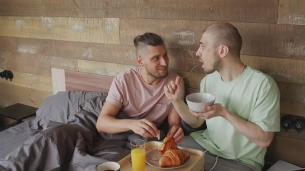 朝ごはんを食べながらベッドに座っておしゃべりする若い同い年の男たちの手持ち撮影 — ストック動画