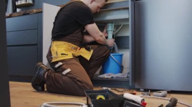 Alet kemeri takmış, mutfak lavabosu dolabının önünde diz çökmüş ve lavabo kapanındaki sızıntıyı tamir eden genç bir erkek tesisatçının el kamerasıyla çekilmiş yavaş çekimi.
