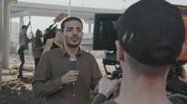 Yavaş çekimde erkek muhabir, çevik kuvvet tam gaz gelmeden önce pankartlı insanları canlı yayında protesto ediyor.
