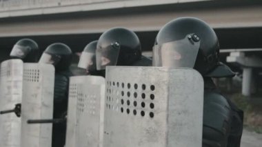 Tanımlanamayan çevik kuvvet polislerinin yavaş çekimleri koruyucu giysiler içinde kalkanları sopalarla dövüyor ve düzen içinde yürüyorlar.