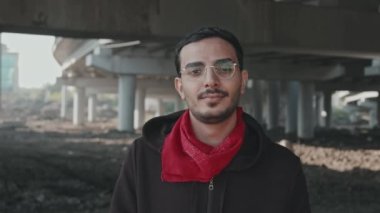 Gözlüklü, köprünün altında kameraya bakan genç ve yavaş çekimde melez bir adamın portresi.