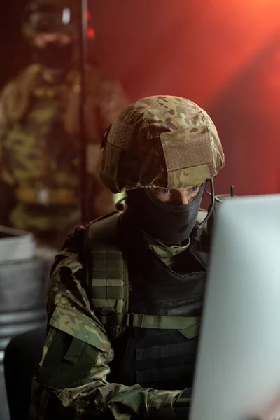Uomo in maschera, casco e uniforme mimetica guardando il monitor del computer — Foto Stock