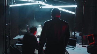 Şapkalı ve deri ceketli bir gencin karanlık bir sığınakta bilgisayarını hackleyen bir kadının dizüstü bilgisayarına doğru yürüdüğü yavaş çekim.