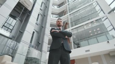 Yüksek modern ofis binasının içinde elleri katlanmış, siyah takım elbiseli profesyonel erkek avukatın düşük açılı ağır ağır portresi.