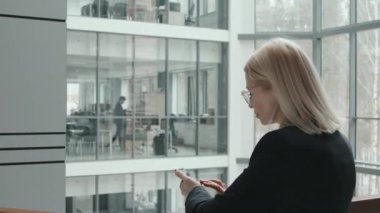 Modern cam ofiste korkuluklara yaslanırken resmi kıyafet giymiş sarışın bir iş kadınının akıllı telefon kullandığı dikiz görüntüsü.
