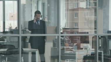 Modern ofis odasında yürürken, siyah takım elbiseli yakışıklı iş adamının ya da avukatın telefon görüşmesi yaptığı orta boy, cam duvarlı, yavaş çekim.